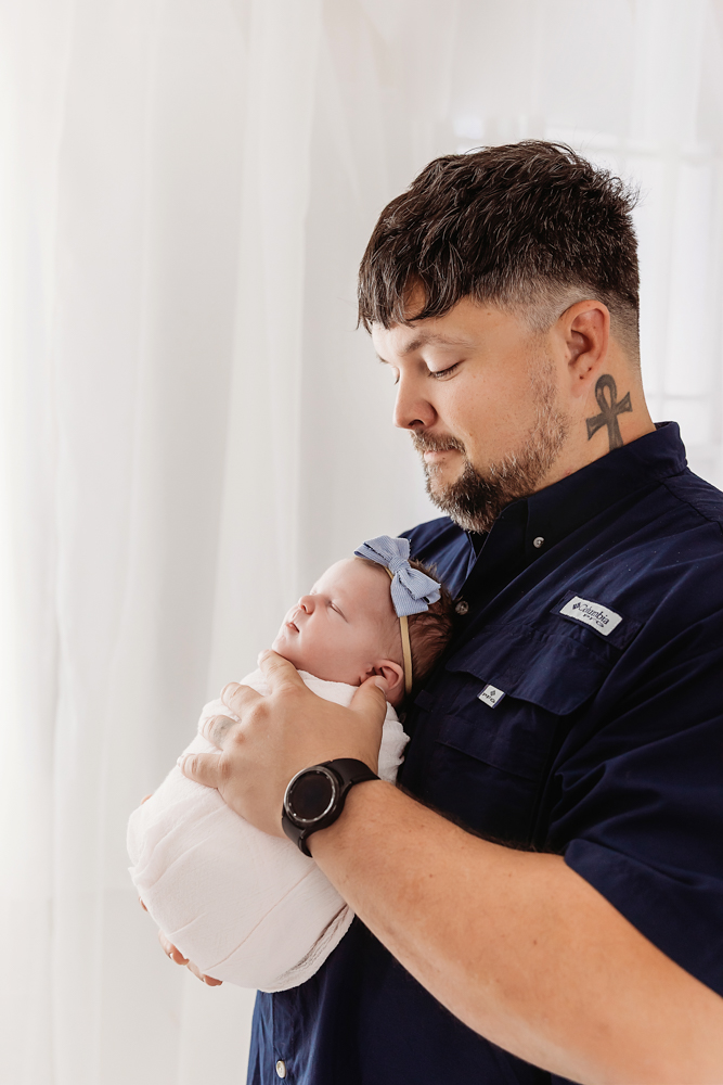 Father holding sleeping newborn baby in white swaddle, wearing navy shirt against soft white backdrop - florence sc photographer