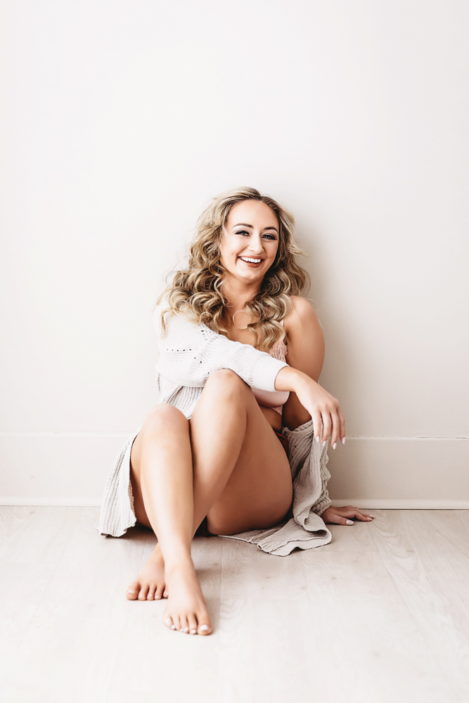 Smiling woman in white knit sweater sitting on floor against neutral wall, soft natural lighting boudoir photography