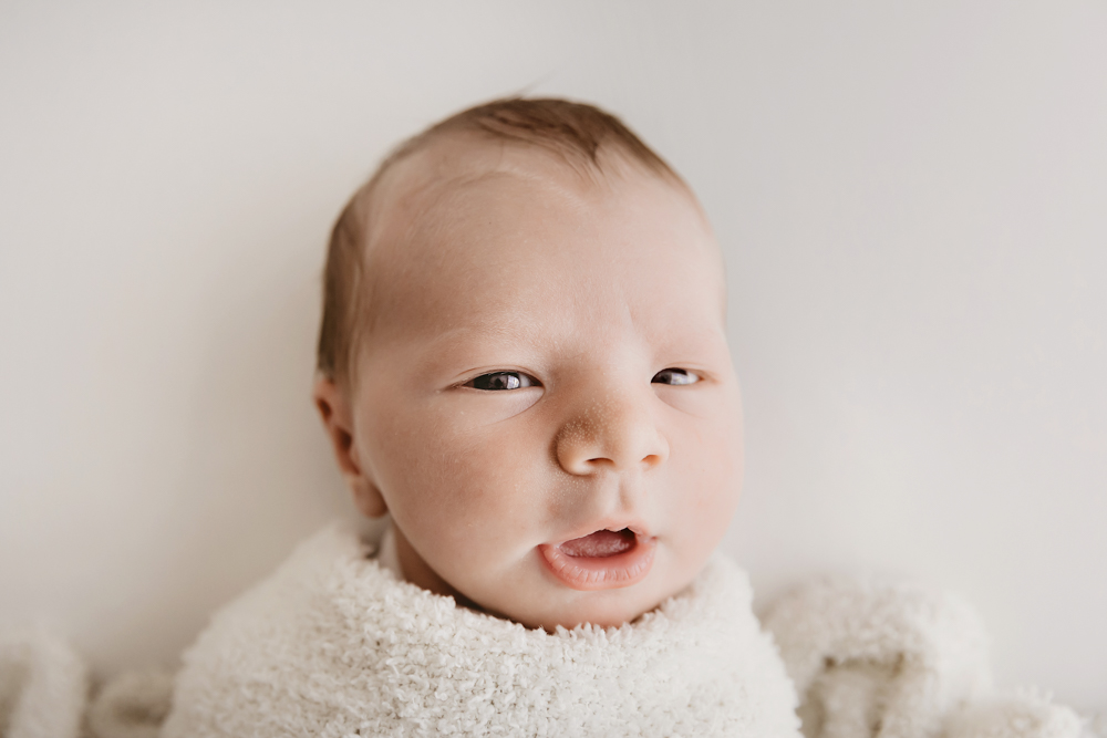 Alert newborn baby with dark hair wrapped in cream textured blanket on neutral backdrop, professional photography