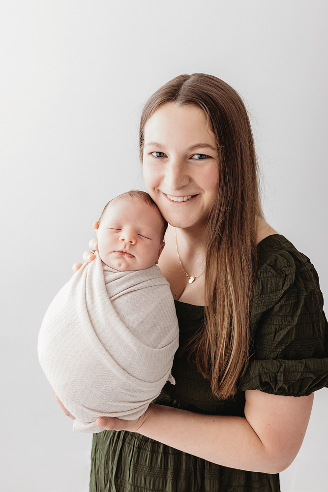Smiling mother in olive green dress holding swaddled newborn in beige wrap, soft studio lighting, florence sc newborn photographer