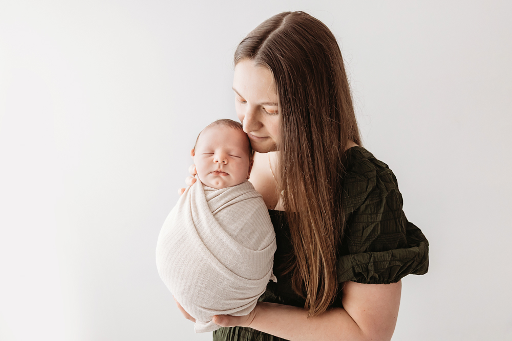 Mother with long brown hair in green top tenderly holding swaddled newborn in cream wrap on white backdrop - florence sc photographer