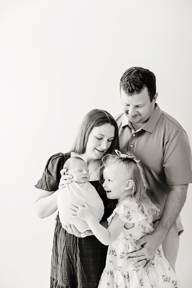 Family of four embracing in black and white studio portrait - parents with newborn and toddler, professional photography near me