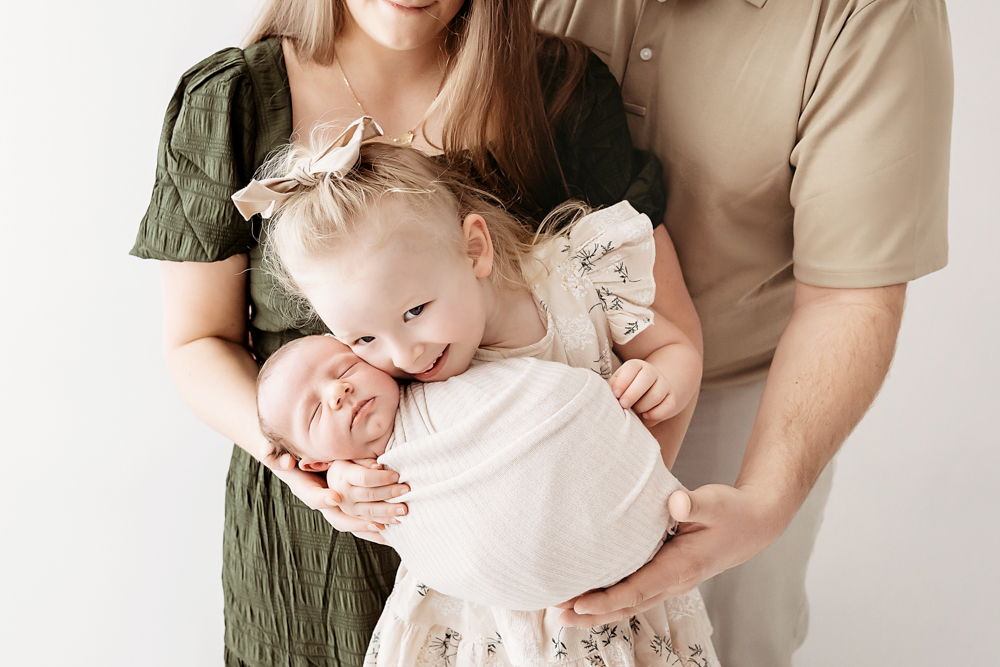 Family portrait at professional photography studio: toddler in floral dress holding newborn baby, with parents in olive green and beige on white backdrop