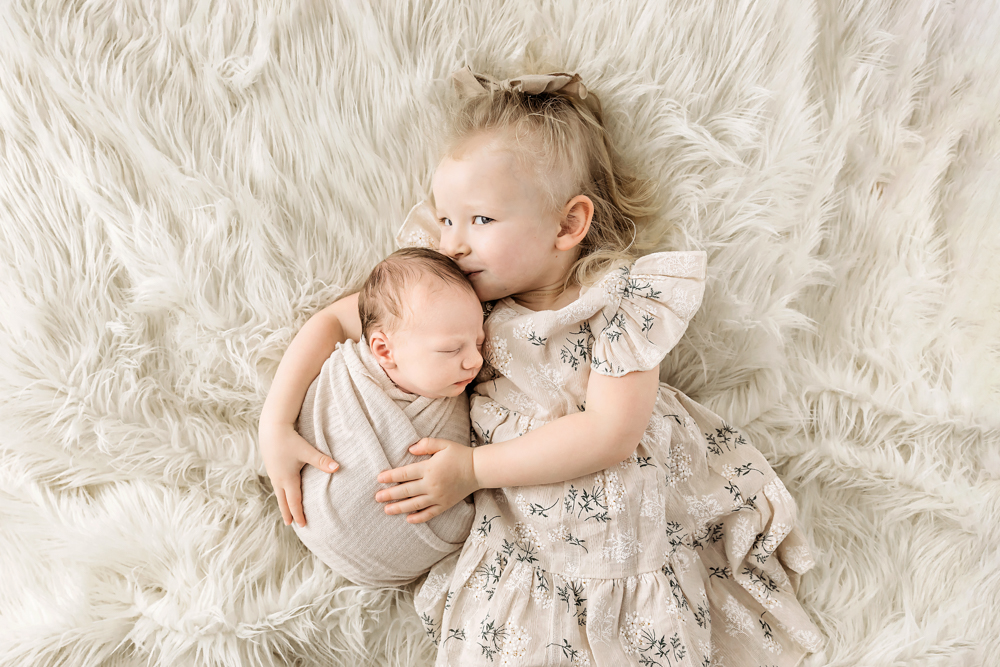 Toddler girl cuddling newborn baby on cream fluffy blanket, professional photography with soft natural lighting