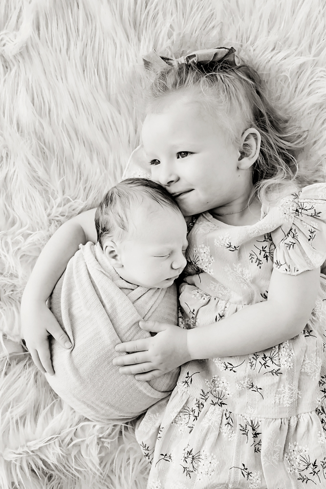 Young child cuddling newborn baby on white fluffy fur backdrop, black and white portrait by professional photographer