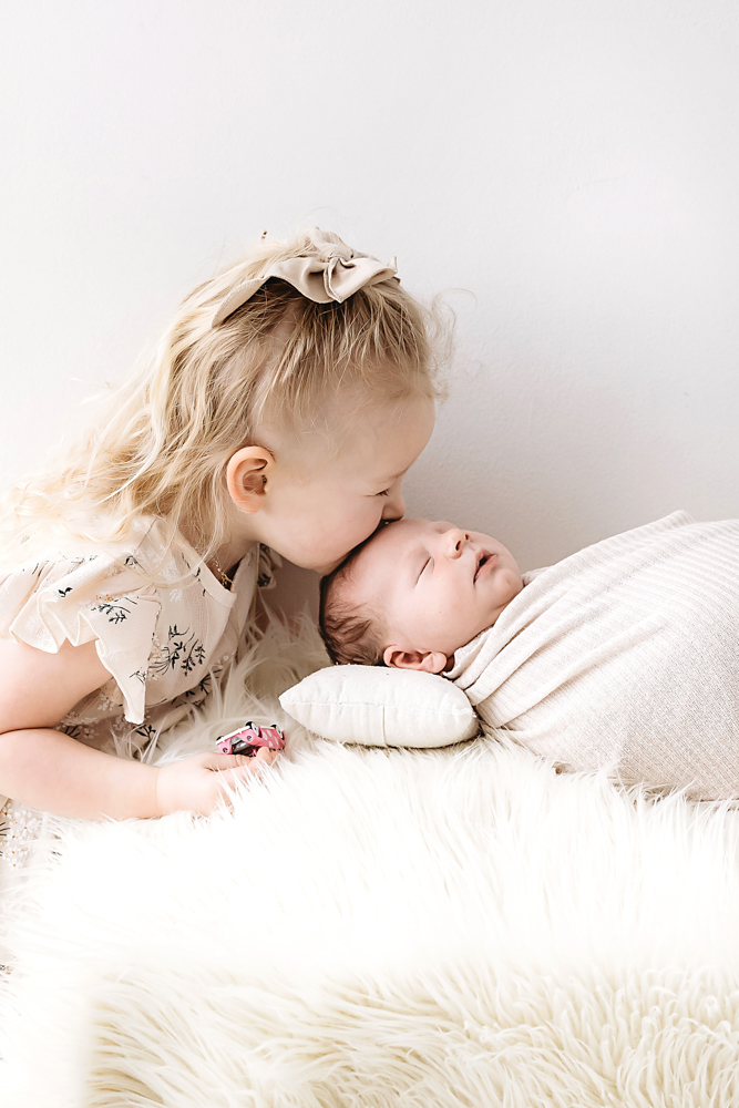 Toddler with blonde hair and bow gently kissing newborn baby on cream blankets - professional newborn pictures Florence SC