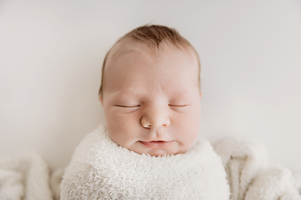 Peaceful sleeping newborn baby wrapped in white textured blanket on cream backdrop by professional photographer