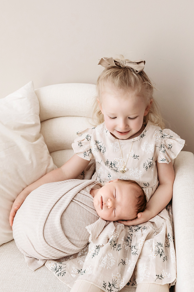 Smiling toddler in floral dress holding sleeping newborn in cream chair - soft natural light newborn photographer portrait