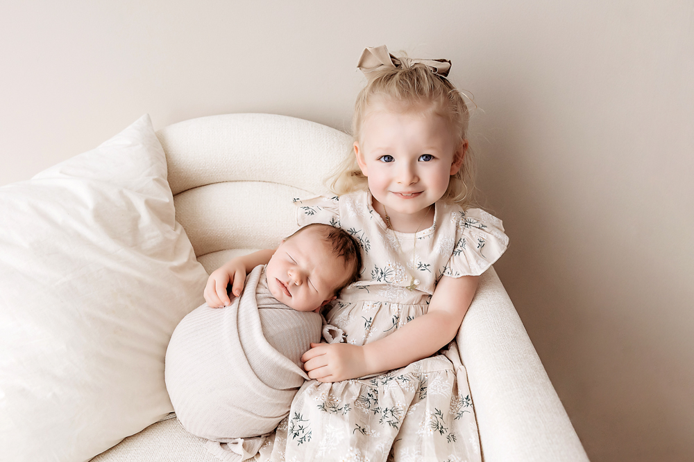 Smiling toddler in floral dress holding sleeping newborn on cream chair, soft natural lighting - Reflection Images by Tracy Rowell