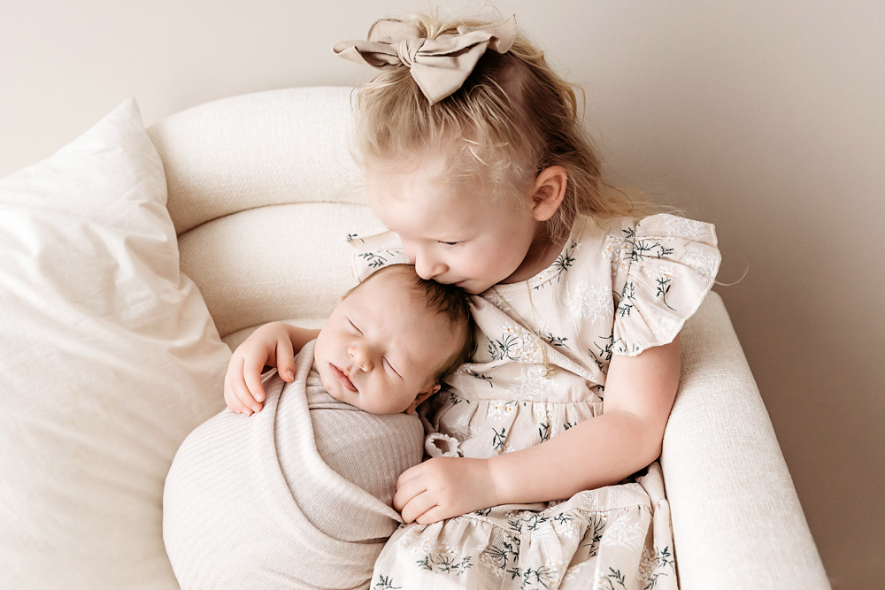 Toddler girl in floral dress with bow headband tenderly holding sleeping baby brother on cream chair - Tracy Rowell