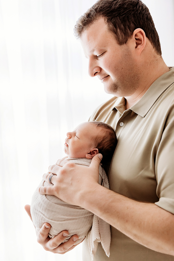 Father tenderly holding sleeping newborn baby in beige wrap, soft natural lighting, newborn photos session in Florence SC
