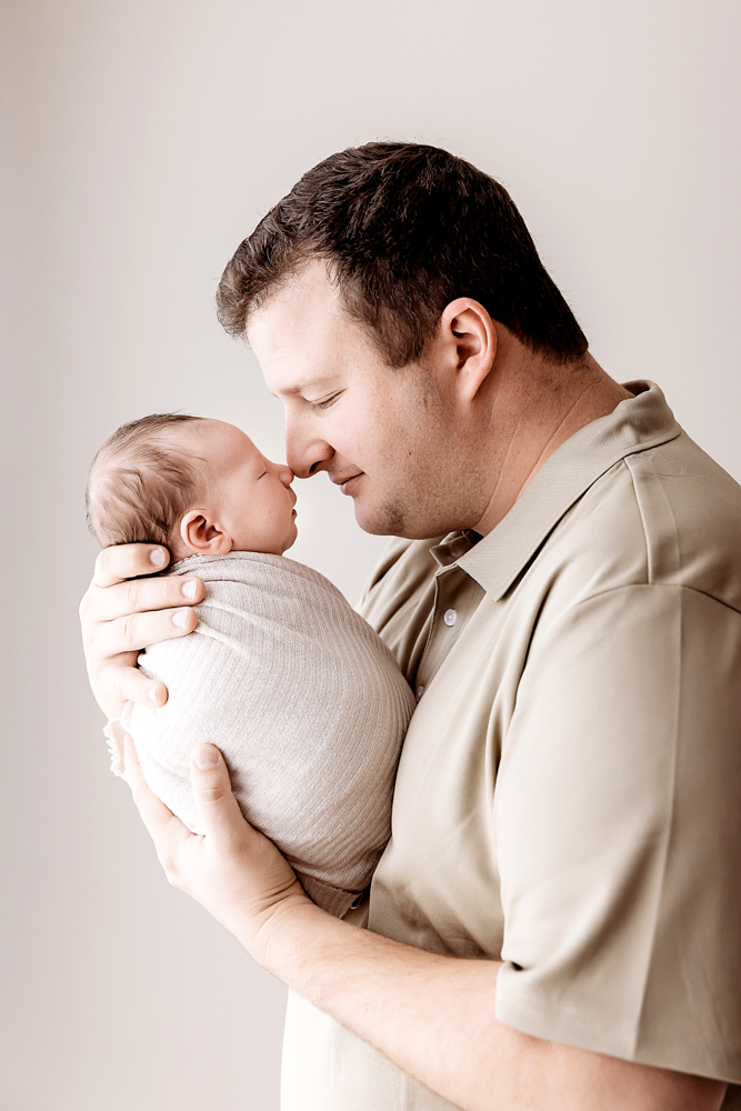 Father tenderly holding swaddled baby in beige wrap, wearing tan shirt, soft lighting for newborn pictures portrait session