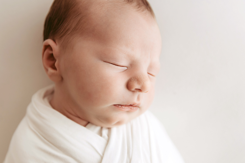 Sleeping newborn baby wrapped in white blanket on cream backdrop, soft natural lighting, professional photography studio near me