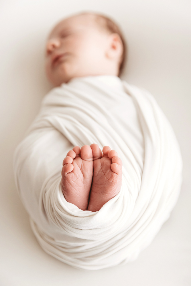 Sleeping newborn baby wrapped in cream blanket on white backdrop, tiny feet visible, soft natural lighting, professional photography near me