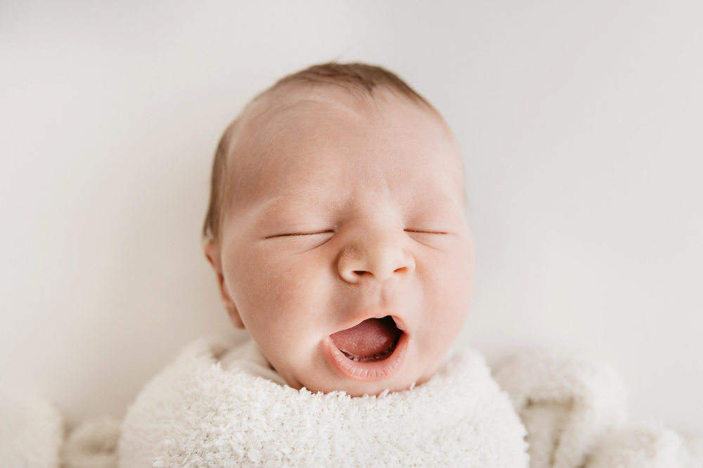 Yawning newborn baby wrapped in white textured blanket on cream backdrop - professional newborn pictures with soft lighting