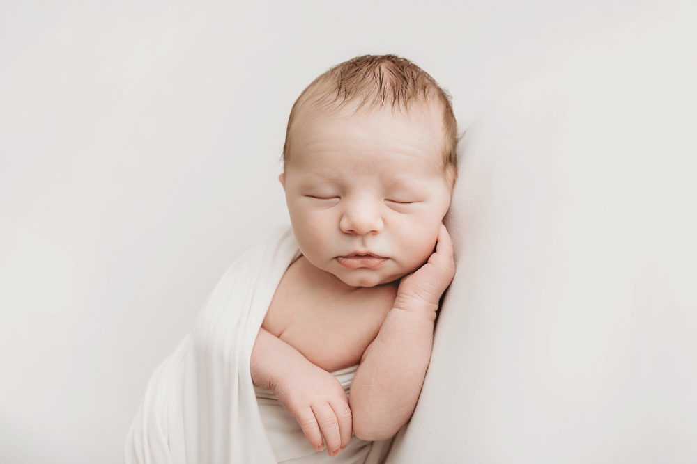 Sleeping baby with hand on cheek on white backdrop - professional newborn pictures with soft natural lighting
