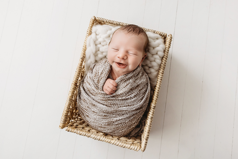 Smiling newborn baby wrapped in beige knit blanket lying in wicker basket with white backdrop - newborn photography session