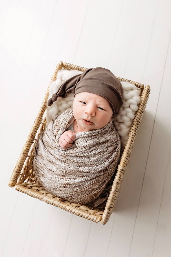 Alert newborn in brown knit wrap and hat lying in wicker basket, soft natural lighting, newborn photographer portrait