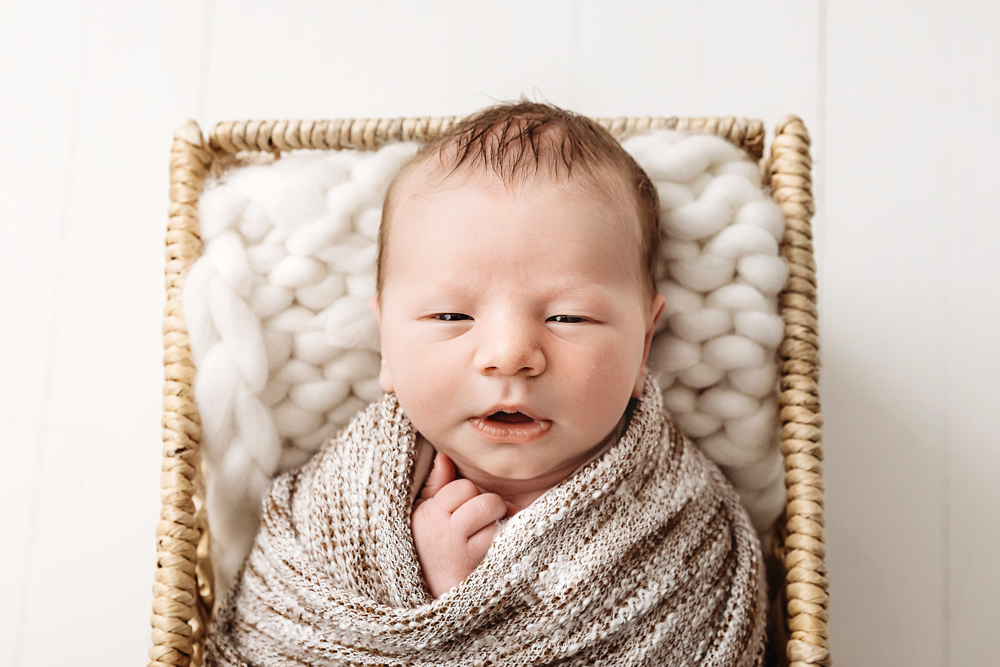 Alert newborn in beige knit wrap lying in wicker basket with white chunky blanket, Reflection Images by Tracy Rowell