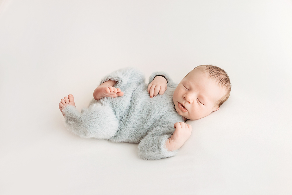 Sleeping newborn baby in soft blue fuzzy outfit on cream backdrop, peaceful pose by Tracy Rowell