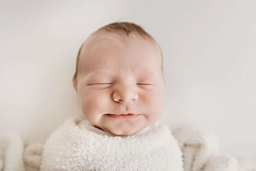 Smiling newborn baby wrapped in cream knit blanket on soft white backdrop, Florence SC newborn photography session
