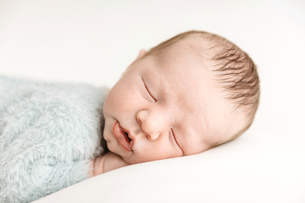 Sleeping newborn baby wrapped in soft blue knit blanket on cream backdrop, professional photographer near me portrait
