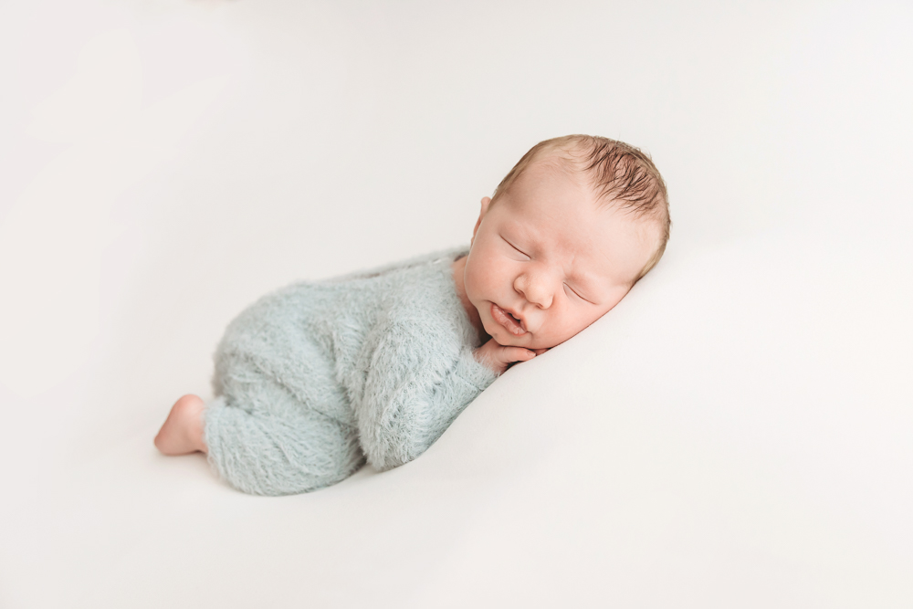 Sleeping newborn baby in sage green knit wrap on white blanket, professional photography studio portrait with soft lighting