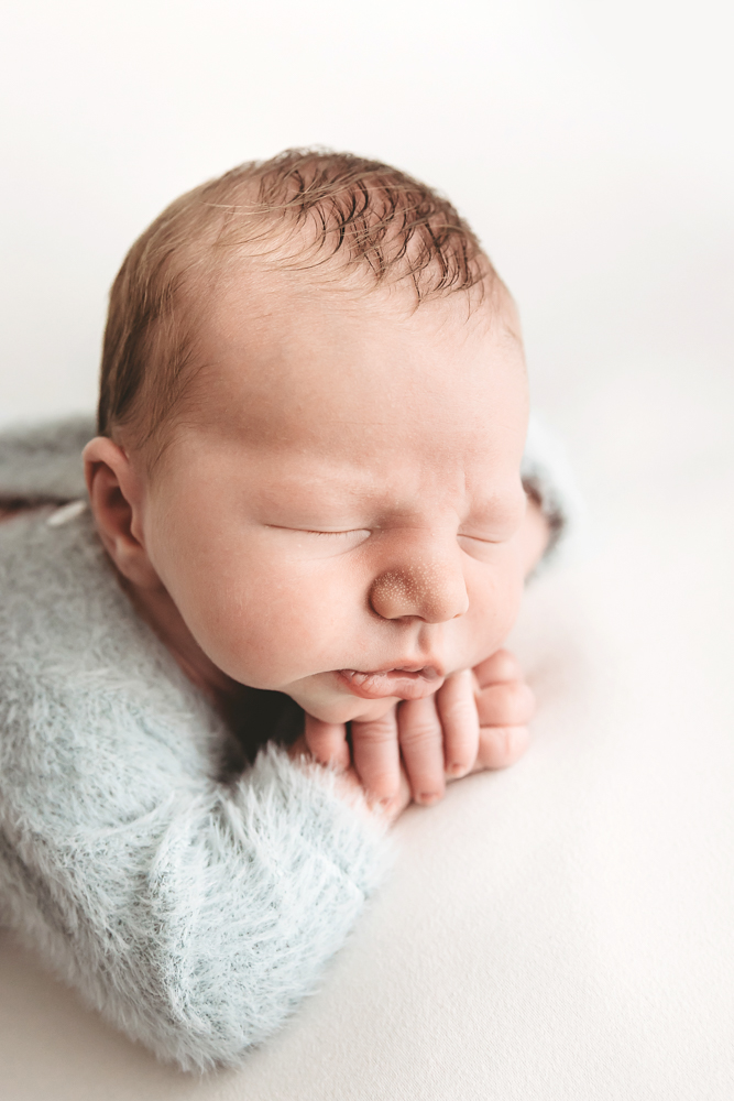 Sleeping newborn baby wrapped in soft gray blanket with hands near face, professional photography on cream backdrop