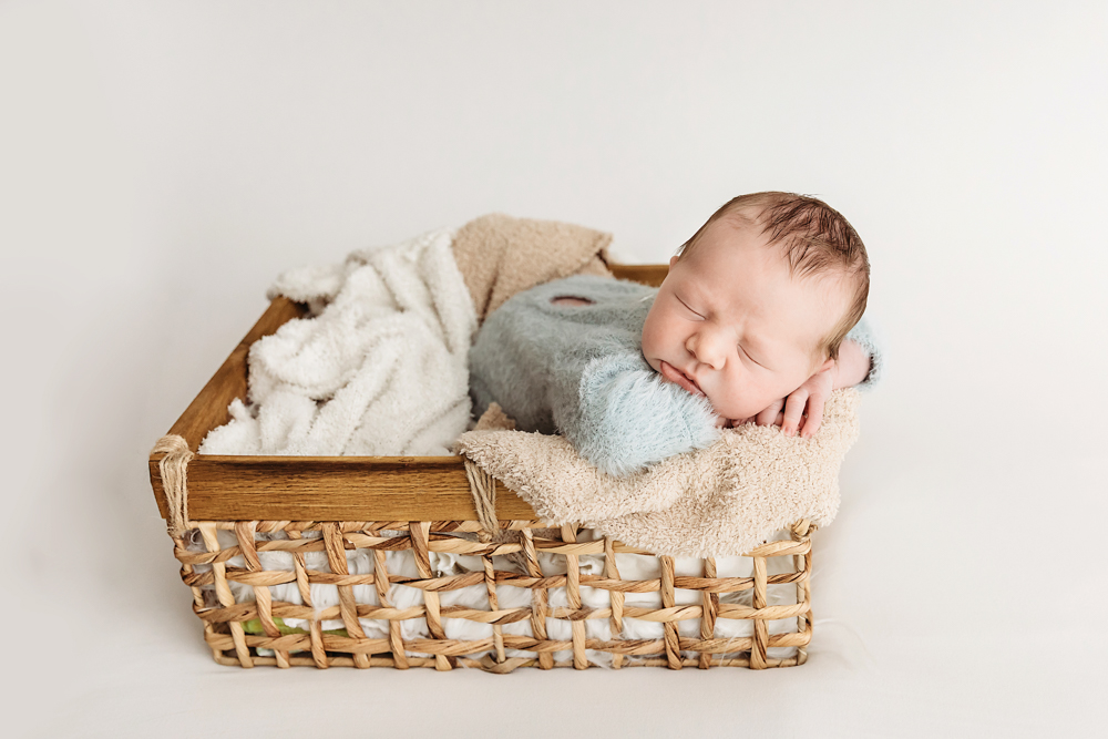 Sleeping newborn in sage green wrap nestled in woven basket with cream blankets, professional newborn pictures studio setup