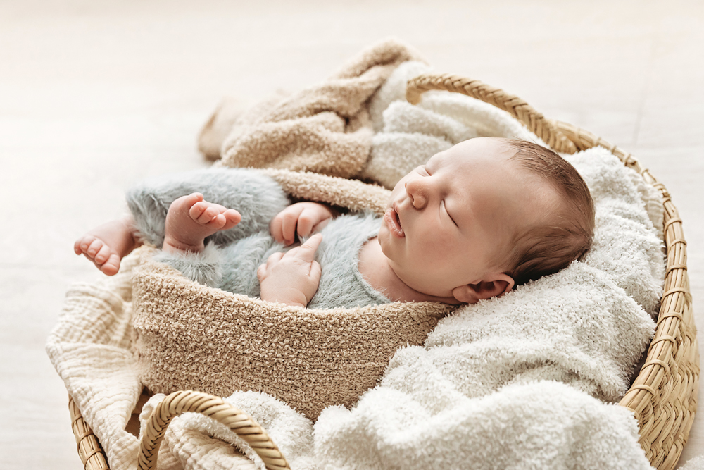 Sleeping baby in sage green wrap nestled in wicker basket with cream blankets, soft natural lighting newborn photography