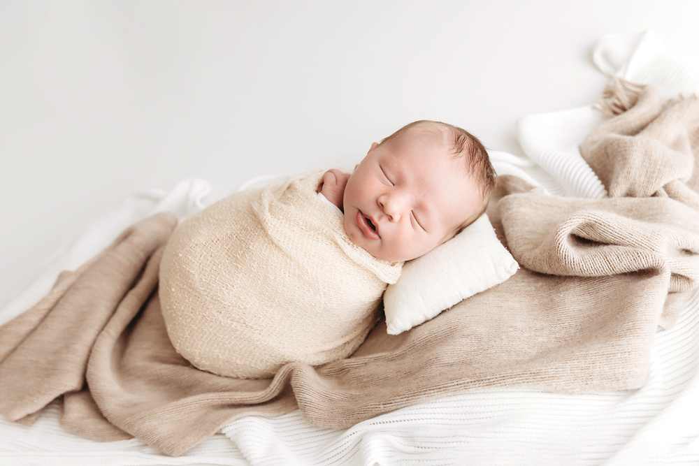Sleeping newborn wrapped in cream knit blanket on soft beige textiles, peaceful expression, newborn photographer portrait
