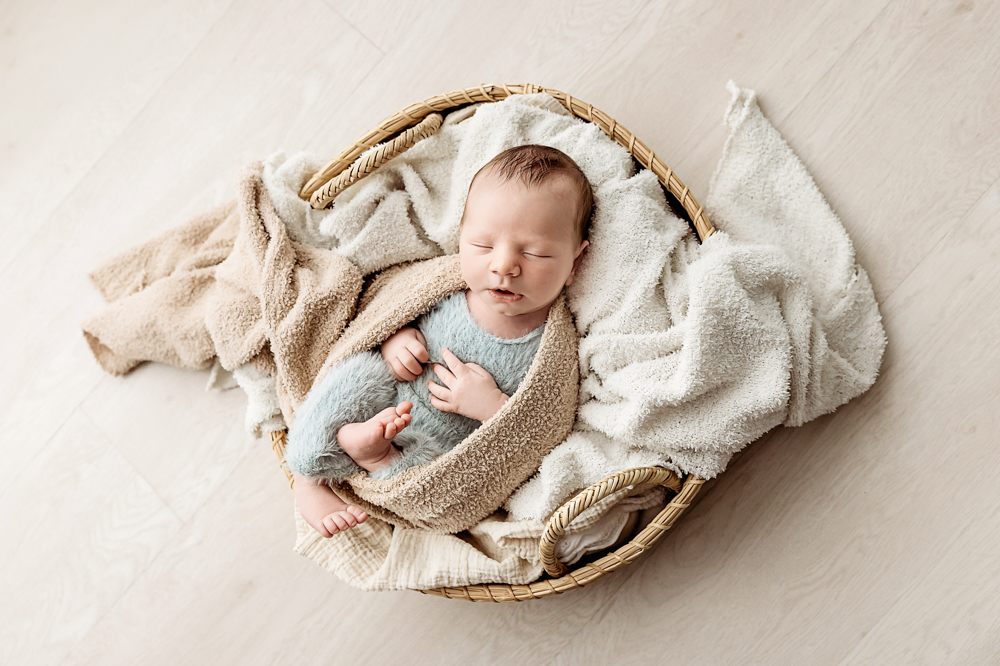 Sleeping newborn in mint wrap nestled in wicker basket with cream blankets, soft natural lighting by newborn photographer
