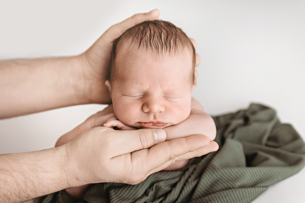 Sleeping newborn cradled in adult hands on white backdrop with olive green textured blanket by Tracy Rowell