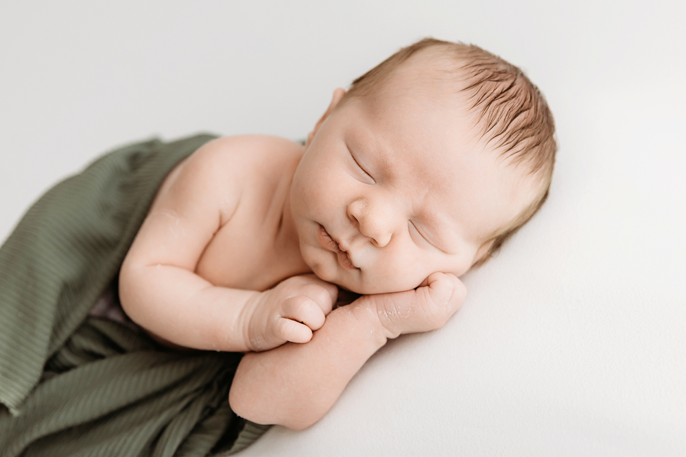 Peaceful sleeping baby wrapped in sage green fabric on cream backdrop for newborn photos with soft natural lighting