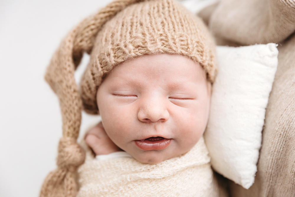 Peaceful newborn baby sleeping in beige knit hat and wrap on cream textured blankets, soft natural lighting - Reflection Images by Tracy Rowell