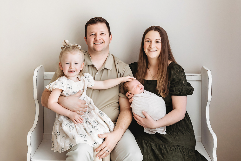 Happy family of four on white bench: dad in beige, mom in green dress holding newborn, toddler in floral dress - Tracy Rowell