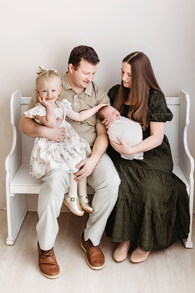 Family newborn photos on white bench - parents in olive green dress and beige shirt holding sleeping baby and toddler girl