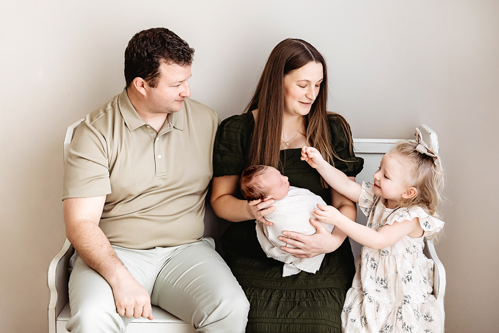 Family of four sitting on white bench for newborn pictures - parents in green dress and beige polo holding baby in white
