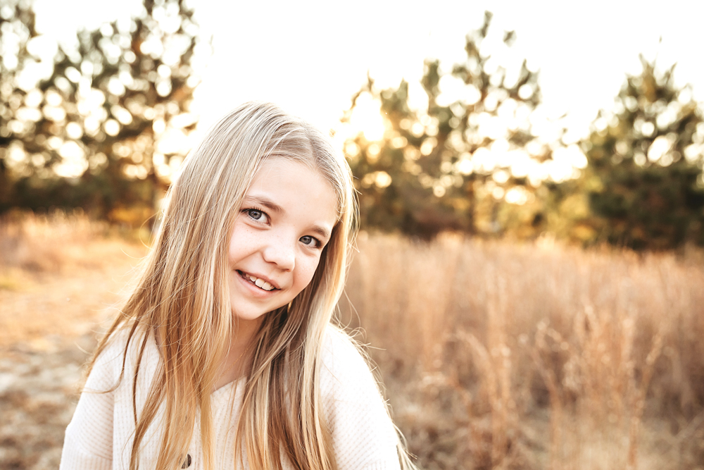 Smiling teen with long blonde hair in white sweater during golden hour fall family pictures in field with trees