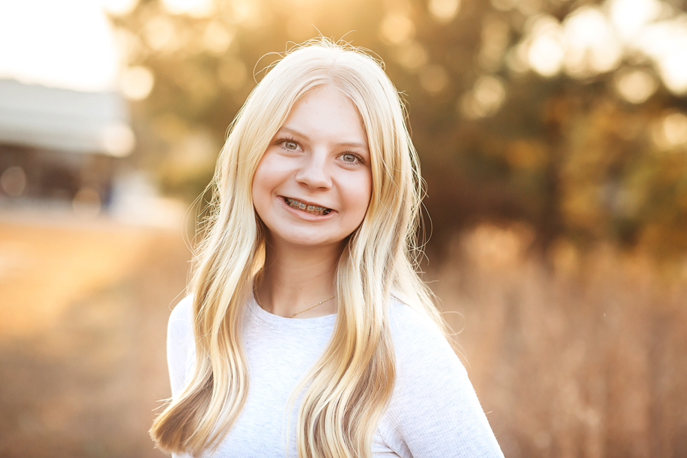 Smiling blonde teen with braces in white top, golden hour lighting, blurred autumn background - florence sc fall family photographer