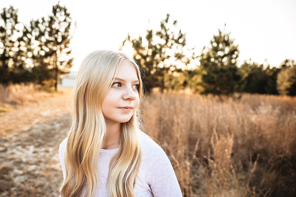 Teen with long blonde hair in white sweater gazing thoughtfully in golden hour field, professional photography studio near me