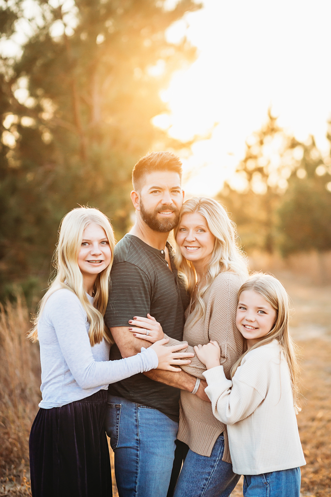 Family of four embracing outdoors at golden hour, professional photographer captured warm sunset lighting through trees
