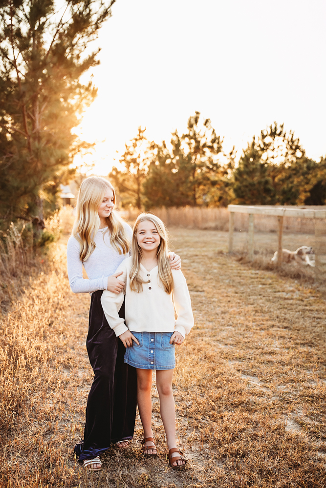 Teen and child sisters embracing in golden hour light, cream sweaters and denim, fall family photographer session outdoors