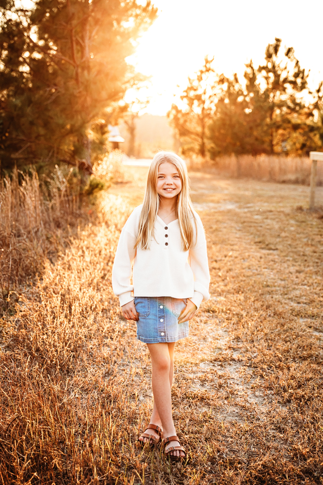 Smiling child in white top and denim skirt standing on golden dirt path at sunset, Tracy Rowell portrait photography