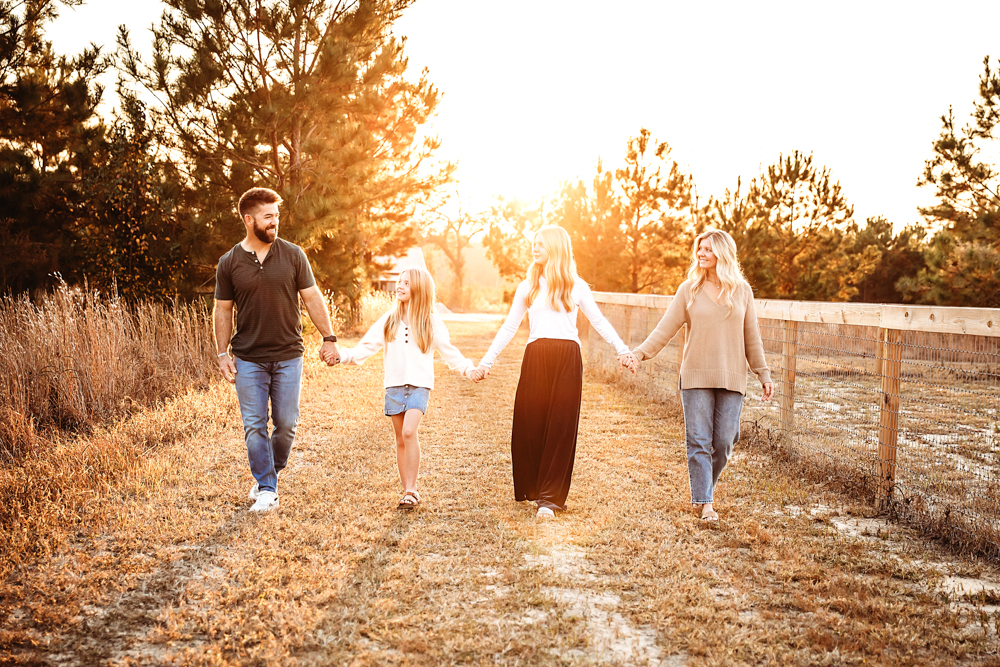 Family of four holding hands walking through golden field during fall family photos with warm sunset lighting and rustic fence