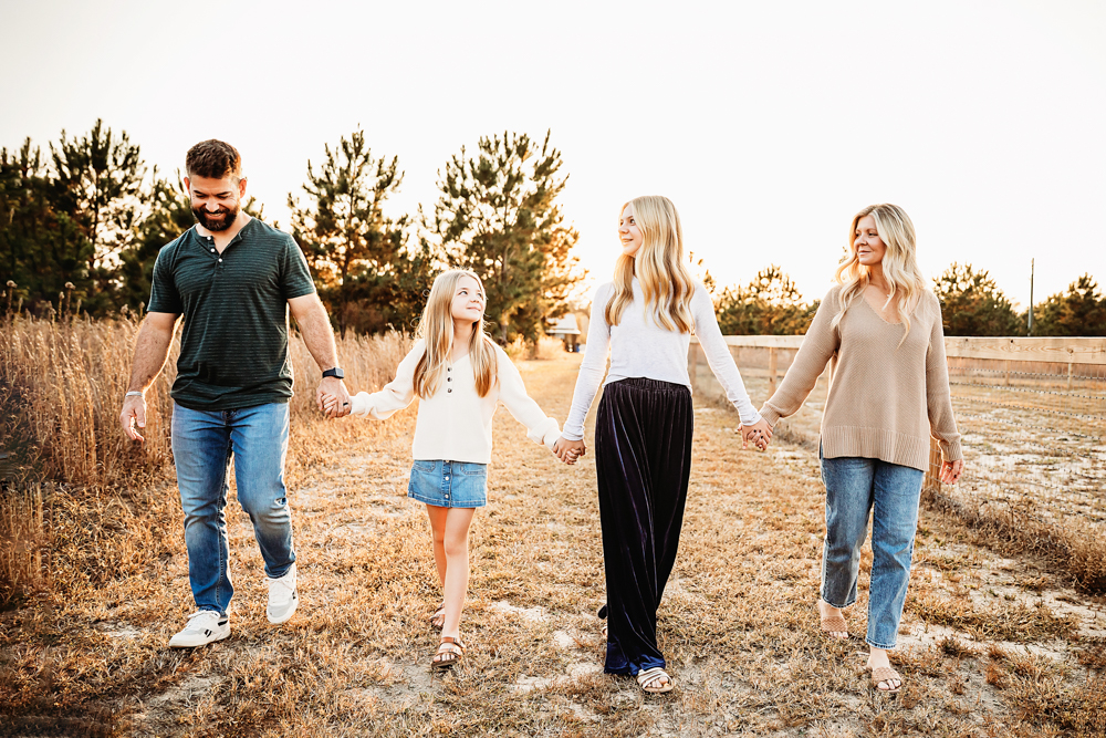 Family of four holding hands walking in golden field at sunset for fall family pictures, wearing casual earth tones
