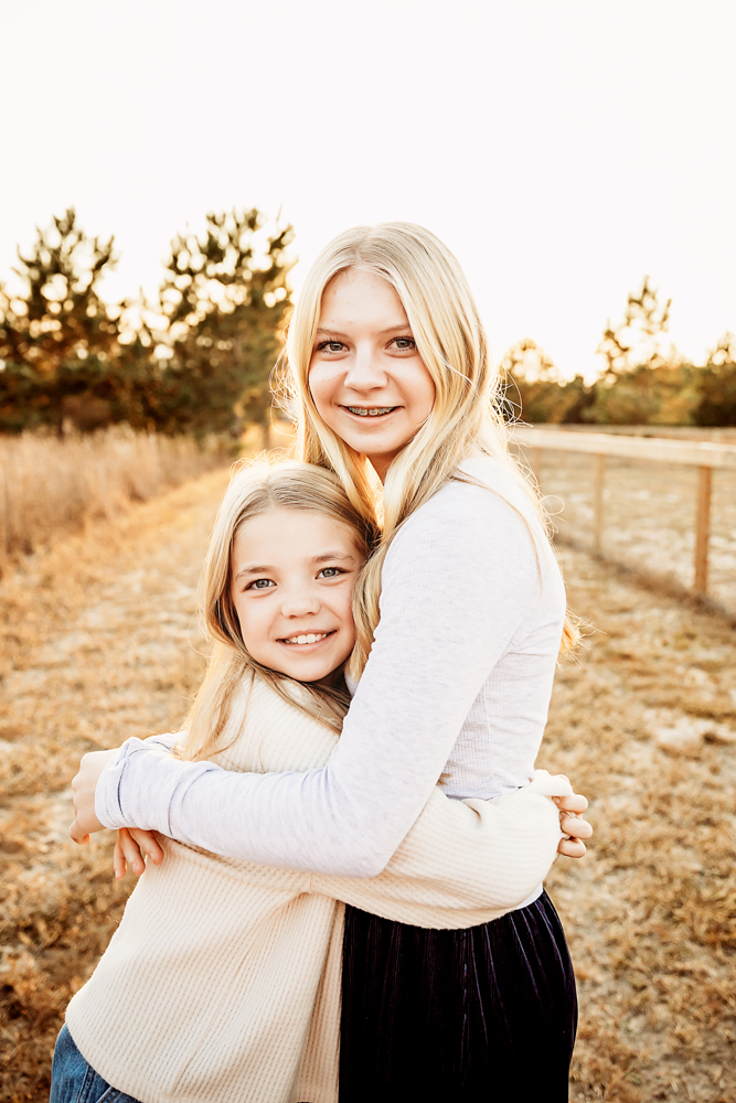 Two blonde sisters hugging and smiling in golden hour light, wearing cream sweaters in rural field - florence sc photographer