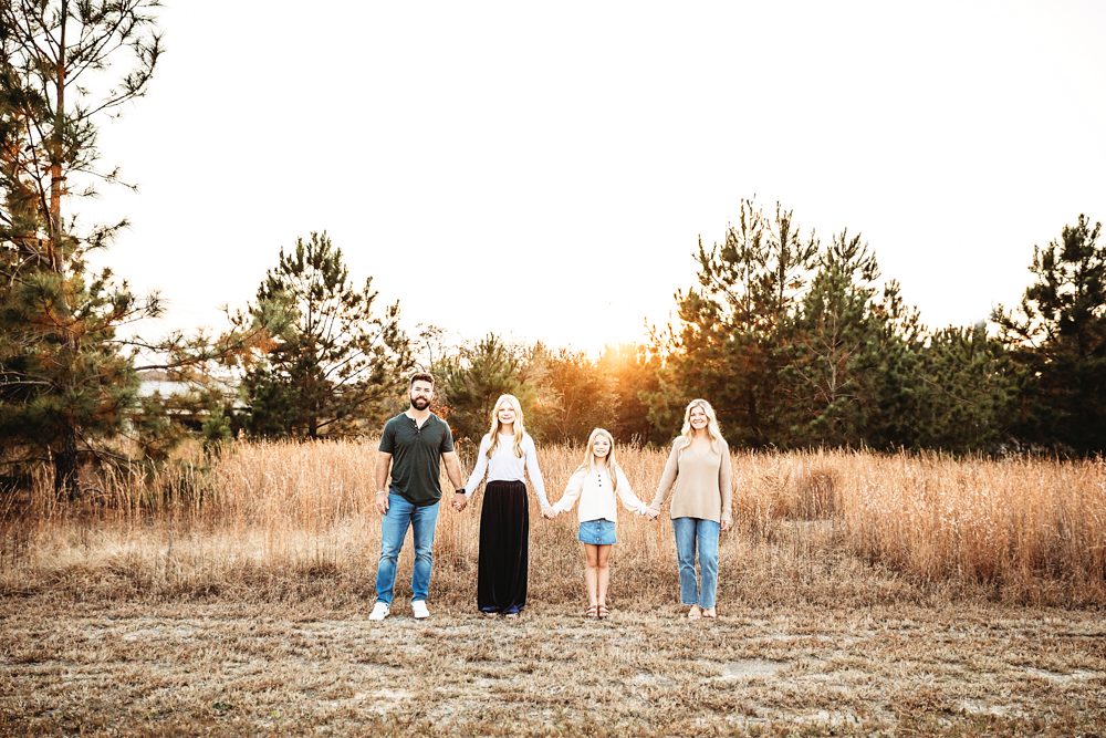 Family holding hands in golden field at sunset, adult man in black shirt, women in white and beige, child in blue - professional photography near me