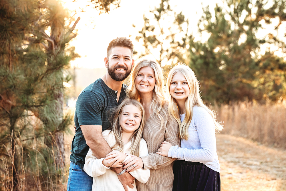 Family of four embracing outdoors at golden hour, professional photographer captured warm lighting through trees backdrop
