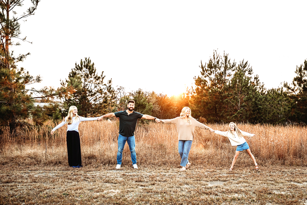 Family of four holding hands in golden field at sunset, professional photographer near me captures warm outdoor portrait